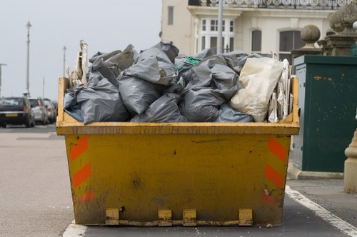 A large yellow industrial skip filled with numerous black and white rubbish bags, positioned on a street near residential buildings in Morden. The bags are made of plastic, some are tightly packed and crumpled, and others appear loosely filled. The skip shows signs of dirt and wear, with visible rust and grime marks along its base and sides. The skip is placed on pavement, close to the curb, with a few cars visible parked along the street behind it. In the background, there are residential buildings with white facades, bay windows, and decorative stone elements, indicating a typical urban suburban setting. The sky appears overcast, contributing to a neutral, diffuse lighting that highlights the textures of the rubbish and the skip surfaces. The scene reflects a rubbish removal activity, consistent with services offered by Office Clearance Morden, operating in the local area.
