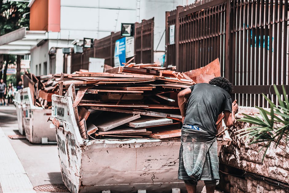 A worker dressed in a green uniform and safety helmet is standing on the side of a street, operating or inspecting a large, industrial rubbish collection vehicle. The vehicle is positioned on an aspha