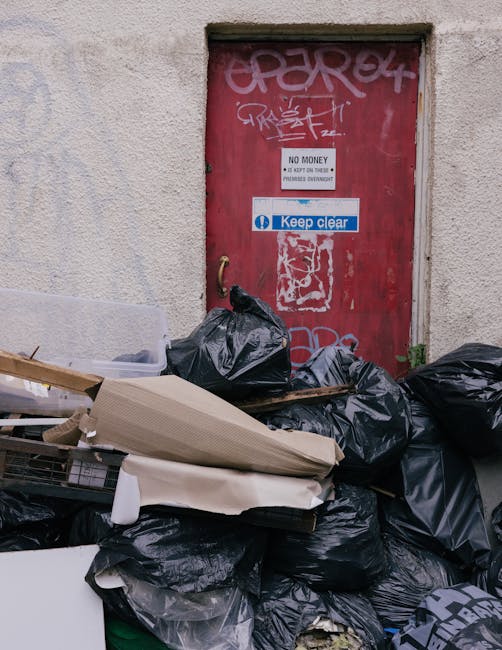 Two large black plastic rubbish bags, slightly crumpled and leaning against each other on a concrete pavement, are positioned near the edge of a street. The bags appear to be filled with household was