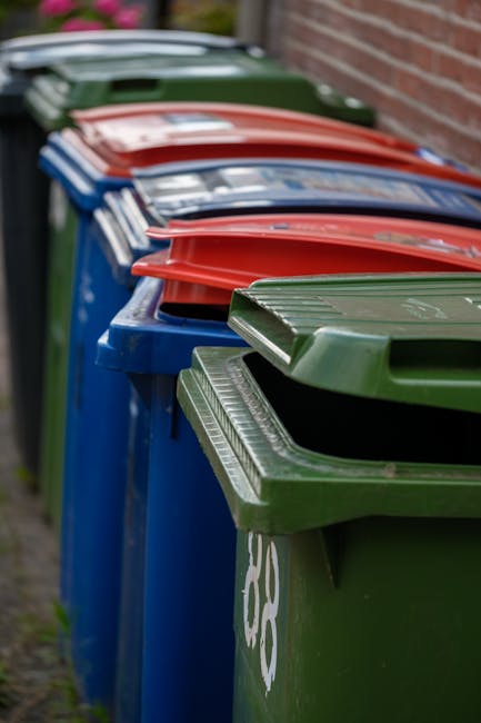 A row of large at-capacity wheelie bins made of durable plastic, positioned along a pavement with their lids closed. The bins are painted in different colors including green, blue, red, and black, eac