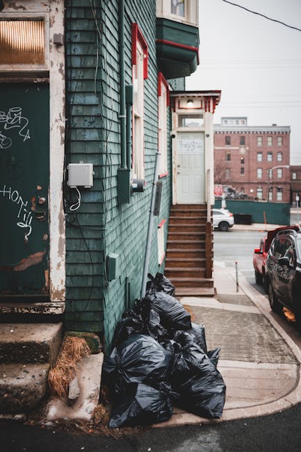 A sanitation worker wearing a fluorescent yellow safety vest and red long-sleeved shirt is standing at the rear of a large red rubbish collection truck, which is parked on the side of a street. The tr