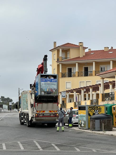 A sanitation worker dressed in green protective clothing and a helmet is standing on a paved urban street, operating a large waste collection vehicle with its rear hopper open. The vehicle appears to