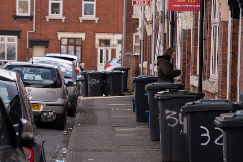 A residential street scene showing a row of dark grey wheelie bins placed along the sidewalk outside brick terraced houses with white-framed windows and stone decorative sills. The bins are lined up i