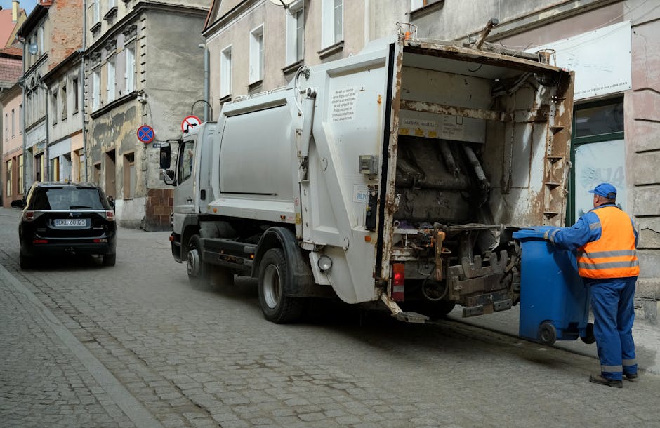 A residential street scene showing a row of dark grey wheelie bins placed along the sidewalk outside brick terraced houses with white-framed windows and stone decorative sills. The bins are lined up i
