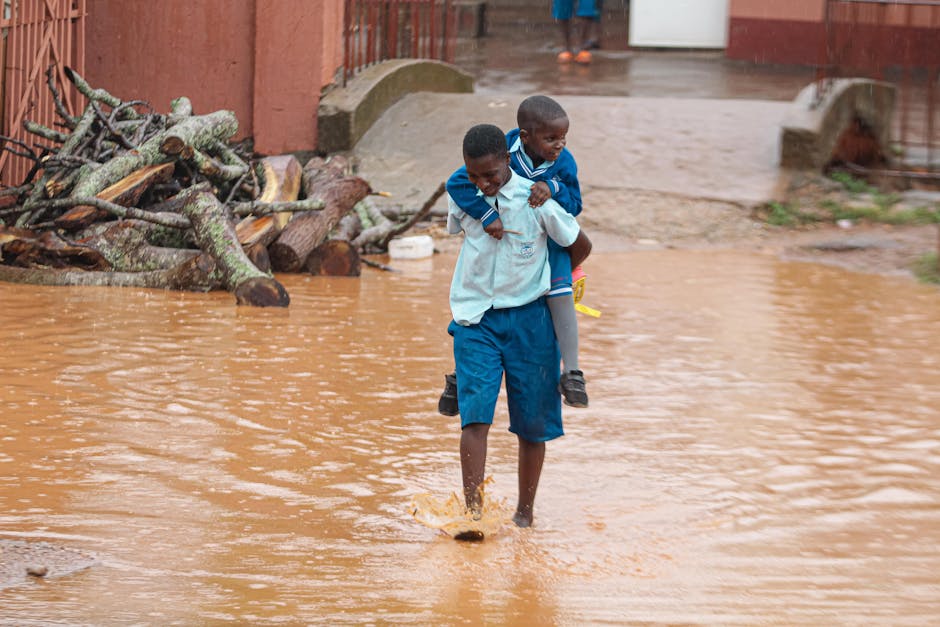 A street in an urban residential area is flooded with muddy water reaching halfway up the lower floors of multicolored buildings. The buildings feature various architectural styles with facades in sha